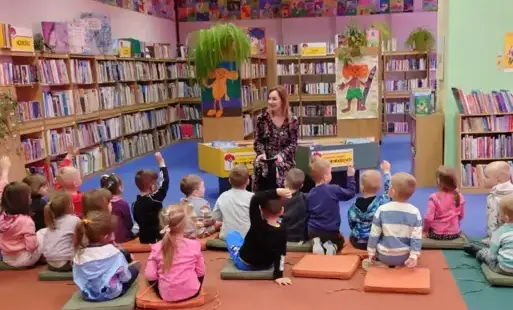 Dzieci siedzą na kolorowych poduchach na dywanie przed bibliotekarką i biorą udział w pogadance. Niektóre z nich podnoszą rączki do odpowiedzi. W tle znajdują się regały z książkami.