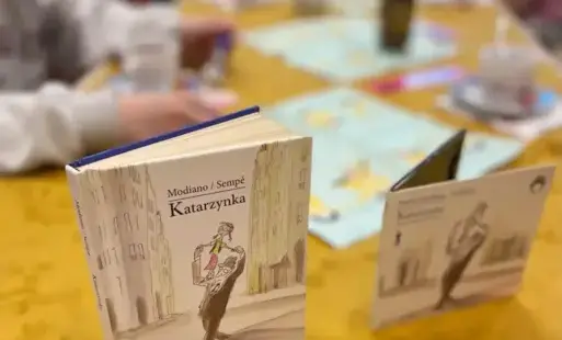 Książka i auiobook o tym samym tytule stoją na nakrytym obrusem stole, w rozmytym tle siedzą osoby.
