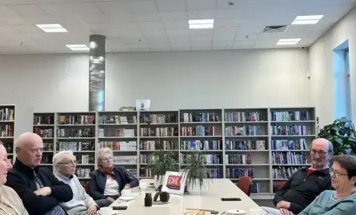 Na zdjęciu na stole stoi książka „Obcy” Alberta Camus. Dalej ustawiona jest ramka z logo Dyskusyjnego Klubu Książki, kwiaty doniczkowe, filiżanki z kawą lub herbatą. Przy stole siedzą też uczestnicy spotkania. Za stołem widać regały z książkami.