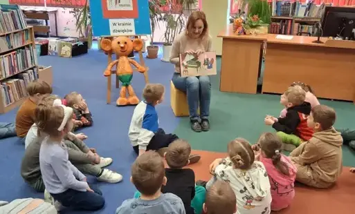 Dzieci siedzą na dywanie, a bibliotekarka pokazuje im obrazek w książce, na którym mały Mozart gra na klawesynie.