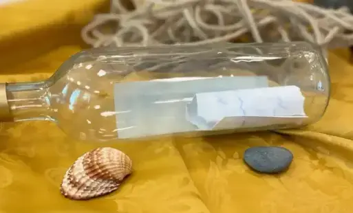 Na udrapowanym obrusie leżą rekwizyty: butelka z listem w środku, muszla, szary kamień.