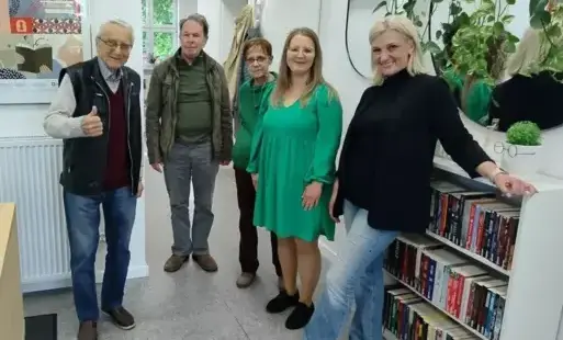 Piątka uczestników spotkania: dwóch mężczyzn od prawej i trzy kobiety od lewej stoi w bibliotece, przy wejściu, przy dwóch lustrach, na które „spływają” zielone liście. Jedna z kobiet opiera się o białą komodę z książkami, która stoi pod lustrami. Za u