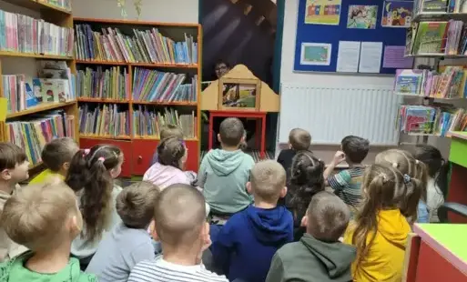 Kobieta za teatrzykiem Kamishibai, przed nią siedzą dzieci, po bokach regały z książkami.