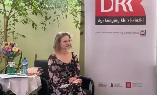 Pisarka, siedzącą przy stoliku, w tle widać rollup z nazwami organizatorów.
