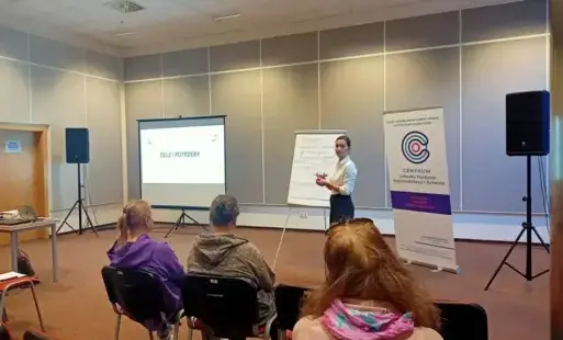 Na pierwszym planie widać sylwetki uczestników siedzących na krzesłach. Dalej, na środku sali, stoi prowadząca spotkanie.  Za nią stoi tablica, na której zapisuje uwagi dot. problematyki warsztatów, a także ekran projekcyjny, na którym widnieje napis „cel
