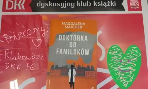 Książka „Doktórka od familoków” Magdaleny Majcher leży na plakacie Dyskusyjnego Klubu Książki. Na górze, nad książką jest logo Dyskusyjnego Klubu Książki oraz Instytutu Książki. Po prawej stronie, przy książce jest zielone serce z namalowanym fioletową