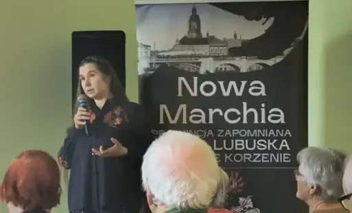 Kobieta stoi z mikorofinem przed publicznością. Za jej plecami baner Nowej Marchii