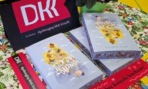 Na stole leżą trzy książki „100 dni słońca” o barwionych brzegach, tabliczka z logo DKK oraz zakładki do książek.