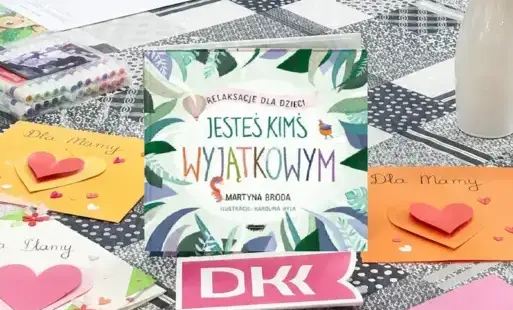 Zdjęcie przedstawia fragment stołu, na którym ustawione jest logo DKK oraz inne elementy, w tym tytułowa książka 'Jesteś kimś wyjątkowym'