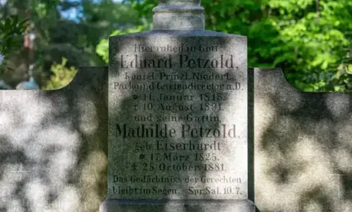 Kamienny, zabytkowy nagrobek z wyrytymi napisami w języku niemieckim, umieszczony na tle zielonej roślinności.