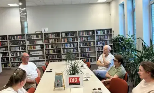 Na zdjęciu na stole stoi książka „Nikt nie odpisuje” Eun-jin Jan. Obok stoi ramka z logo Dyskusyjnego Klubu Książki, za nią kwiat doniczkowy, filiżanki z herbatą. Przy stole siedzą uczestnicy spotkania. Za stołem widać regały z książkami, a po prawej stro