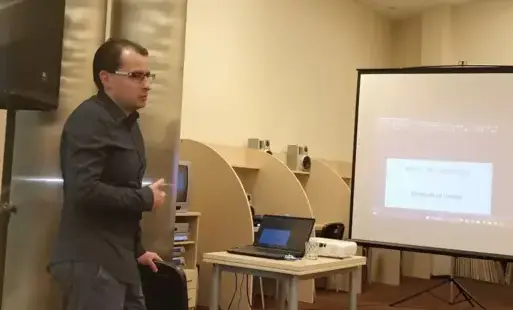 Prowadzący spotkanie mówi do zebranych, koło niego laptop, za nim ekran.