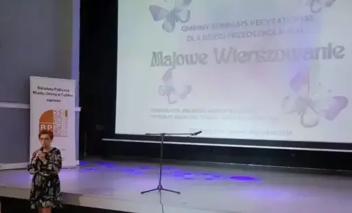 Po lewej stronie sceny stoi kobieta, w tle na ekranie projekcyjnym wyświetlany jest plakat.