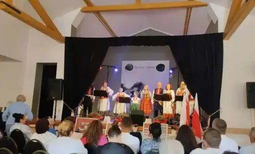 W centrum zdjęcia znajduje się scena, na której występuje zespół Rudniczanka. Scenę okala czarna kurtyna. Przed sceną na krzesełkach siedzą uczestnicy wydarzenia.
