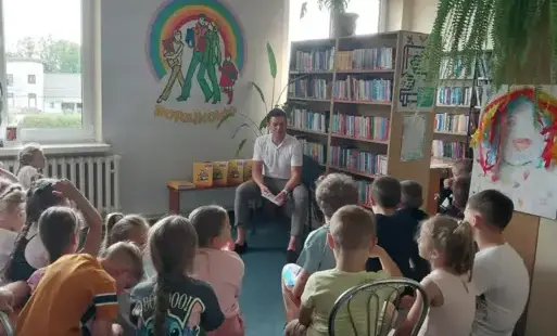 Na jasnej ścianie emblemat biblioteki, regały z kolorowymi książkami, przed tym siedzący mężczyzna trzymający książkę. Przed nim grupa zwróconych w jego kierunku dzieci. Na bokach regałów z książkami plakaty i duża kolorowa praca. W tle zielone rośliny. 