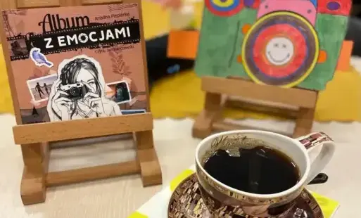 Okładka książki 'Album z emocjami' umieszczona na sztaludze, obok filiżanka kawy oraz druga sztaluga z umieszczonym na niej obrazek z aparatem.
