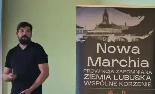 Po prawej stronie baner z hasłem spotkania, po lewej stoi mężczyzna w czarnej koszulce.