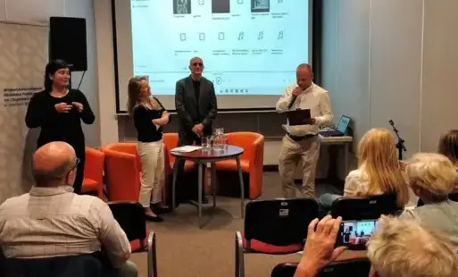 Grupa zaproszonych gości stoi na scenie podczas spotkania w bibliotece. Osoba prowadząca przedstawia ich i oficjalnie rozpoczyna wydarzenie. W tle znajduje się tłumaczka polskiego języka migowego.
