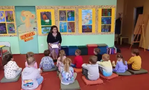 Dzieci siedzą na poduszkach. Przed nimi bibliotekarka czyta książkę. Również siedzi.