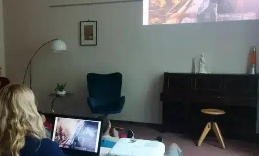 Osoba siedzi przy stole, na którym umieszczony jest laptop. W tle widoczna prezentacja multimedialna na ścianie.