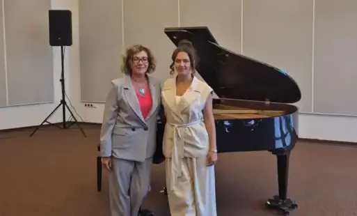 Sala Audytoryjna w gmachu głównym biblioteki. Na tle szarej ściany stoi czarny fortepian, a przed nim na brązowej wykładzinie podłogowej stoi Marina Rusak kierowniczka Sekcji Pianistycznej (po lewej) i Małgorzata Rusak-Mietlińska pełniąca obowiązki dyrekt