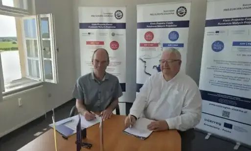 Dwie osoby siedzą przy stoliku, w tle różne plakaty informacyjne