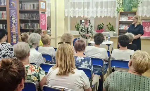Zgromadzona publiczność słucha przemawiającej kobiety stojącej przy stoliku.