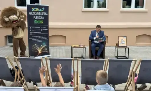 Mężczyzna czyta książkę przez mikrofon, a obok stoi maskotka lwa.