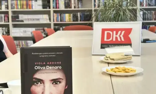 Na pierwszym planie znajduje się stół z egzemplarzem książki „Oliwa Denaro”, dalej logo DKK na tle regałów z książkami. 