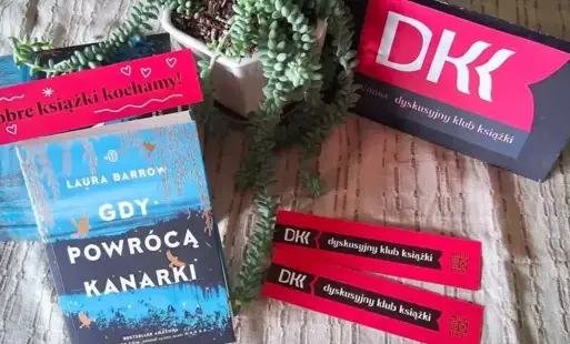 Na stole po lewej stronie leżą omawiane książki. Po prawej stronie jest tabliczka z logo DKK. W środku stoi zielony kwiat doniczkowy.