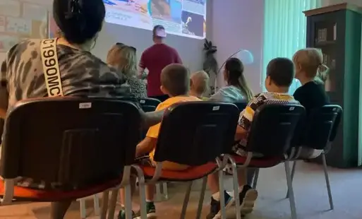 Osoby siedzące na krzesłach, w tle osoba stojąca przy wyświetlanym na ścianie obrazie.