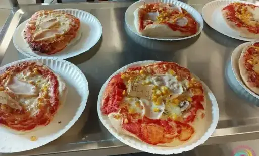 Małe pizze z różnymi dodatkami, ułożone na papierowych talerzykach i oznaczone imionami dzieci.