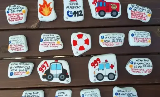 Kilkanaście płaskich kamieni, pomalowanych na biało i ozdobionych kolorowymi rysunkami oraz napisami.