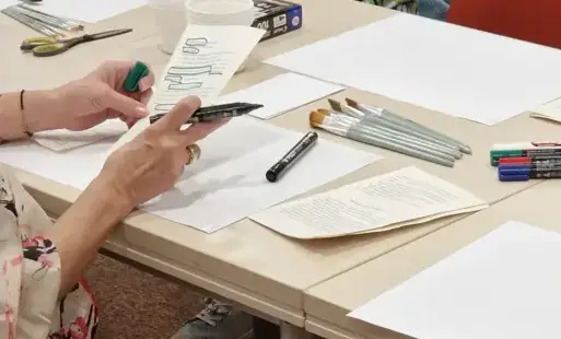 Przy stole warsztatowym siedzą uczestniczki, widać dłonie z flamastrem i kartką z tekstem