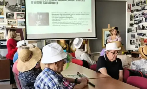Grupa osób siedzi przy stoliku, w tle wyświetlany obraz na ekranie.