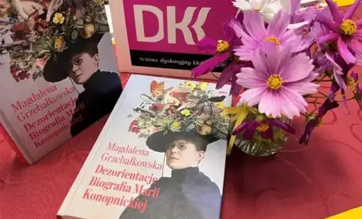 Na stoliku książki, kwiaty w małym dzbanku i logo Dyskusyjnego Klubu Książki