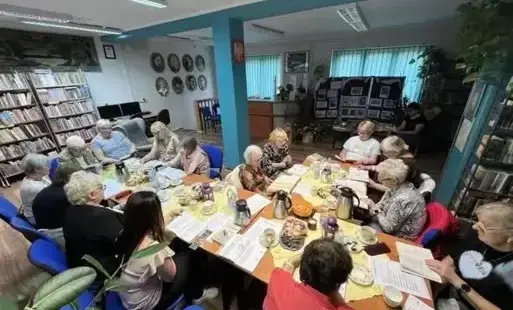 Grupa osób, głównie kobiet, które siedzą wspólnie przy dużym, zastawionym stole w bibliotece i z zaangażowaniem czytają książki.
