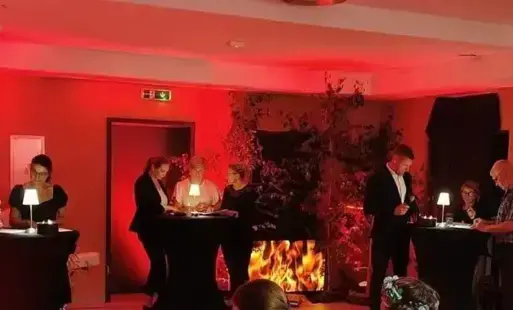 Przy trzech stolikach oświetlonych lampką stoją grupy osób, którzy czytają, na krzesłach siedzą widzowie.