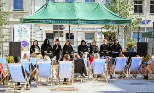 Grupa osób siedzi na leżakach przed sceną, na której występują poprzebierani lektorzy pod zielonym namiotem.