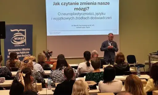 Pomieszczenie biblioteki, na ścianie wyświetlona prezentacja, przy stołach siedzą osoby.