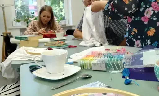 Na zdjęciu widać trójkę uczestników zajęć „Twój T-shirt z charakterem”, dwie kobiety i mężczyznę. Dwie osoby siedzą przy miętowym stole. Prowadząca zajęcia Małgorzata Murowicka-Kozicka stoi.
