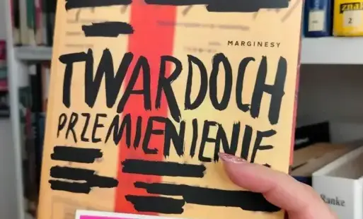 Widać dłoń, która trzyma w górze książkę „Przemienienie” Szczepana Twardocha oraz naklejkę z logiem Dyskusyjnego Klubu Książki. W tle jest biały regał z książkami.