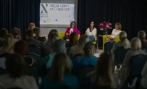 Na scenie, na tle ciemnych zasłon i dużego plakatu z napisem „X Forum Kobiet Aktywnych”, siedzą na fotelach trzy prelegentki prowadzące rozmowę, przed sceną tyłem do obiektywu siedzi liczna publiczność.