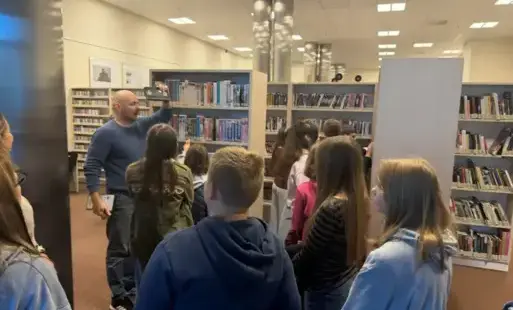 Bibliotekarz trzyma w dłoni oraz prezentuje dzieciom kasetę HVS.