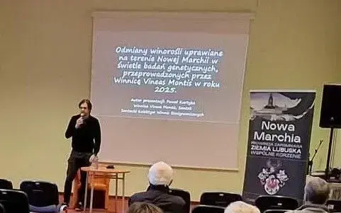 Prelegent z mikrofonem przemawia do publiczności, w tle prezentacja