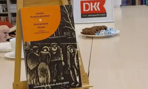 Na stole w bibliotece stoi drewniana sztaluga z książką László Krasznahorkaiego „Szatańskie tango”. W tle widoczne są regały pełne książek oraz znak Dyskusyjnego Klubu Książki. Na stole znajduje się talerz z ciastkami i cukierkami.