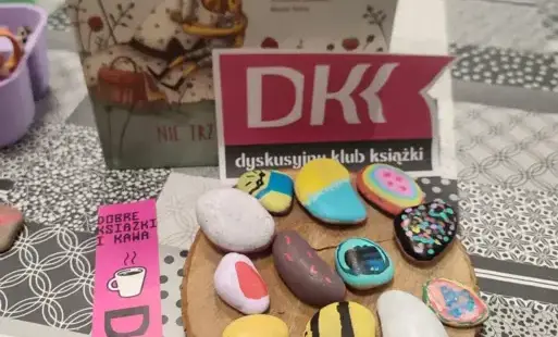 Na stoliku książka, logo i zakładka DKK oraz pomalowane kamienie