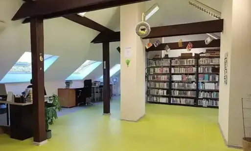Przestronne i jasne wnętrze biblioteki z zieloną podłogą, drewnianymi filarami, regałami pełnymi książek oraz stanowiskami biurowymi pod skośnym dachem.