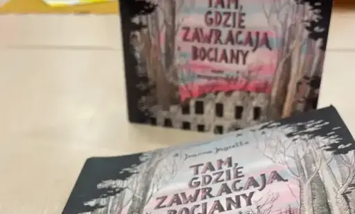 Na stole widoczne opakowanie audiobooka oraz książka o tym samym tytule Tam, gdzie zawracają bociany Joanny Jagiełło, w rozmytym tle filiżanki i siedząca młodzież. 