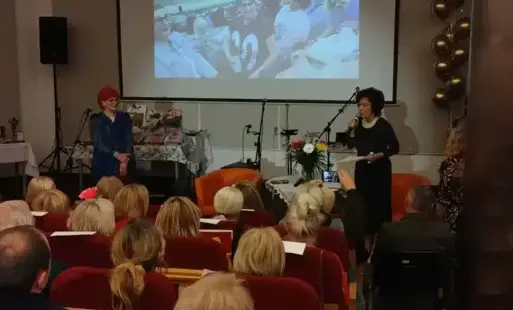 Barbara Schroeder oraz Hanna Kaup przemawiają na scenie, stojąc przed publicznością. W tle prezentowane są zdjęcia przedstawiające działalność Klubu „Na Zapiecku”.